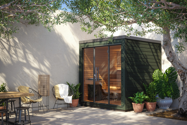 Auroom Mira outdoor sauna with a forest-green exterior, set in a sunny patio with natural greenery and warm wooden interior lighting, showcasing a compact modern wellness design.