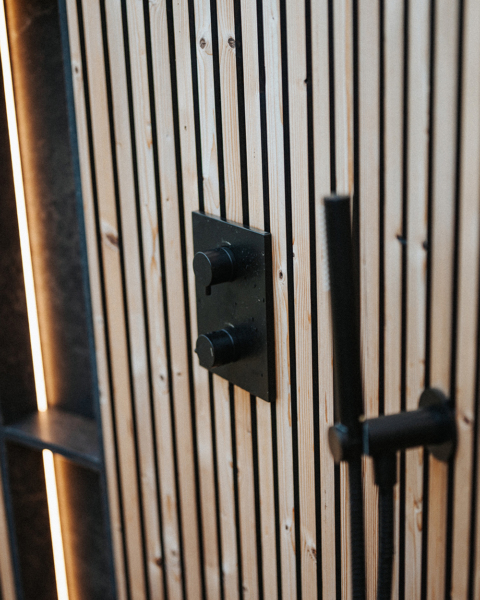 Close-up of a modern wooden wall with vertical slats, featuring a black metal shower control panel and handle.