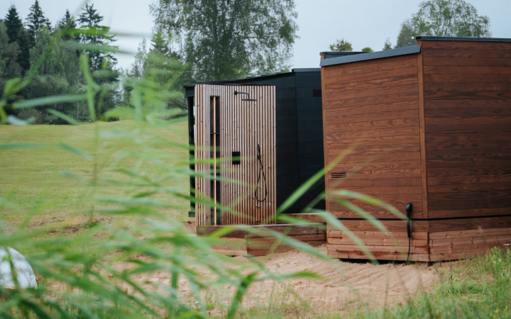 Outdoor wooden shower with a minimalist design, featuring vertical slats and a black showerhead, set against a grassy landscape.