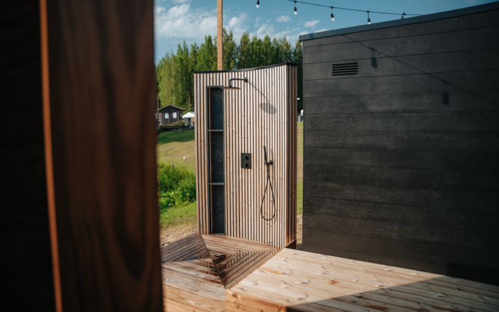 Outdoor wooden shower with vertical slats, a black showerhead, and a platform base, set against a modern black building.