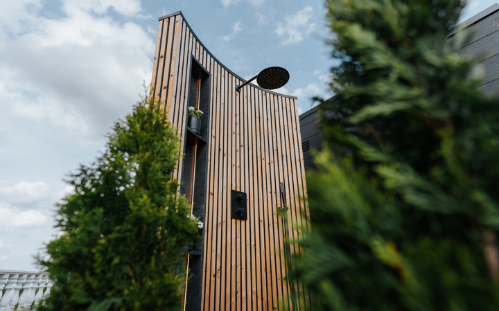 Modern outdoor shower with vertical wooden slats, black fixtures, and small planters, framed by green foliage under a blue sky.