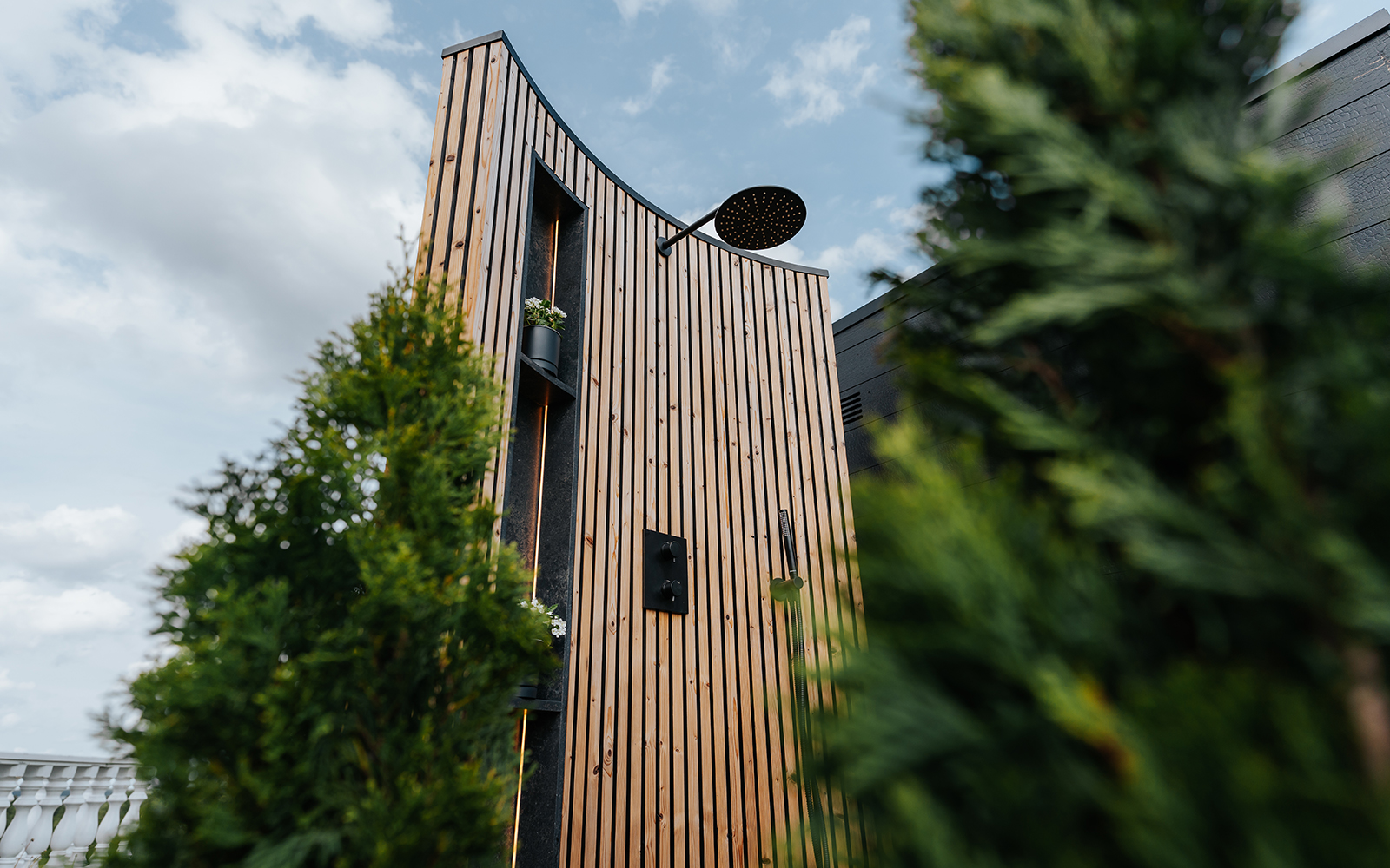 Modern outdoor shower with vertical wooden slats, black fixtures, and small planters, framed by green foliage under a blue sky.