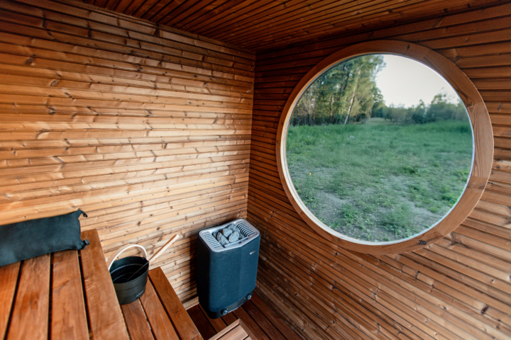 Wooden sauna interior with a round window showing a green field and trees. A bench, a black cushion, a Auroom bucket, and a heater are visible.