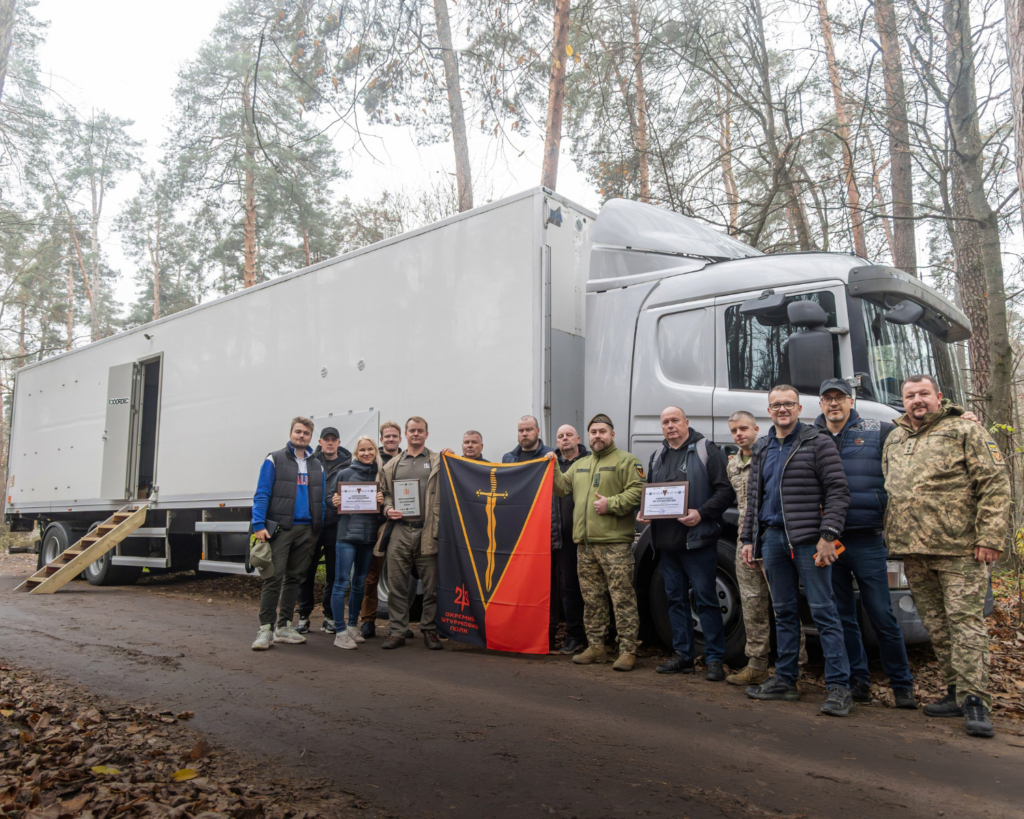 A group of Estonian and Ukrainian team members standing together in front of a large white sauna truck, delivered to Ukraine as part of the Saunas for Ukraine initiative. The team holds certificates and a flag, marking the successful handover of the mobile sauna to the 210th Assault Regiment.
