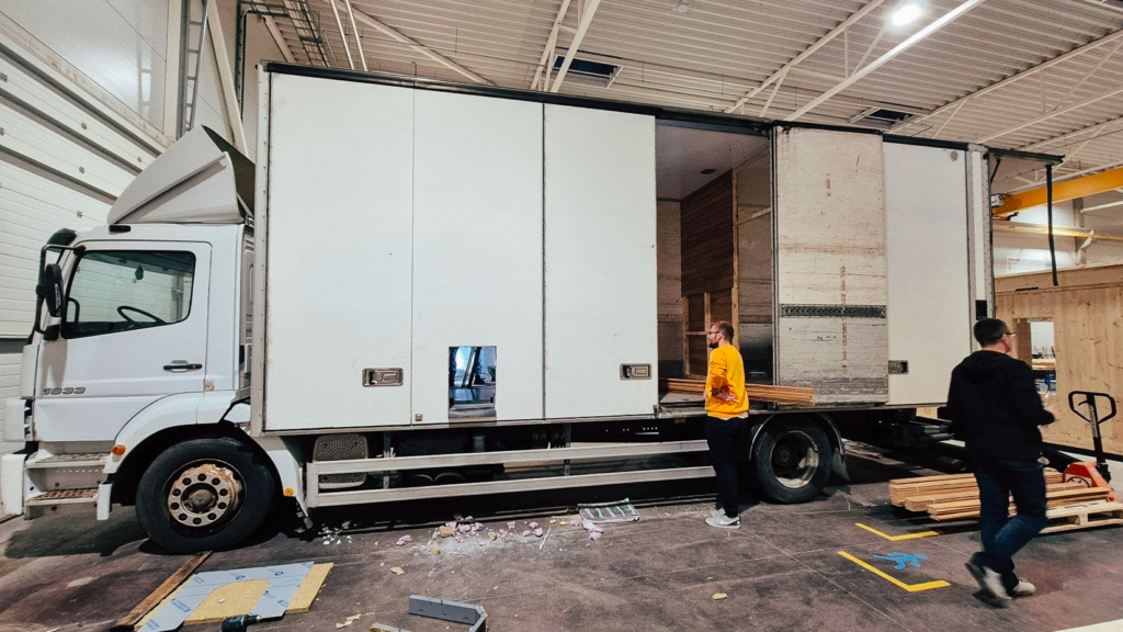 A large white truck being transformed into a mobile sauna at the Auroom workshop. Team members are seen preparing materials and inspecting the structure during the build process.