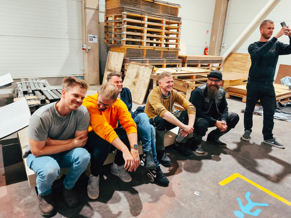 A group of Auroom team members sitting and smiling in the workshop during the mobile sauna construction process. The atmosphere is warm and relaxed after a long evening of teamwork and voluntary effort.